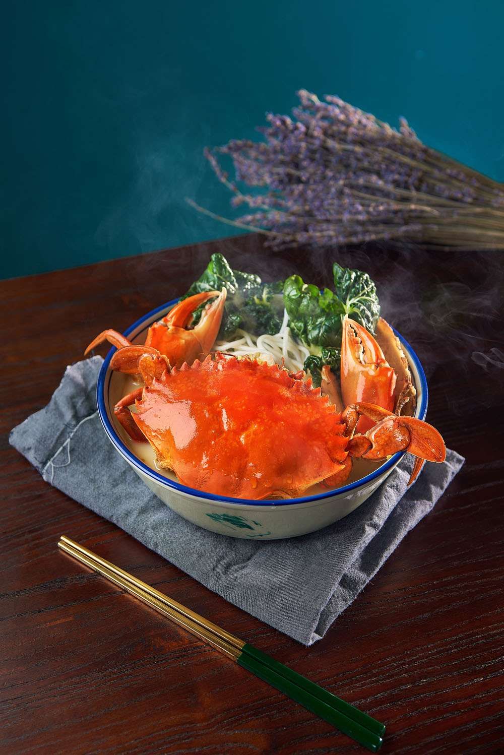 A whole mud crab in a blue-rimmed bowl of bee hoon soup with greens, on a grey cloth.
